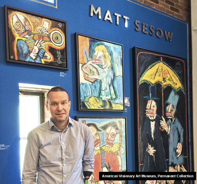 Matt Sesow in front of his paintings on a blue wall at the American Visionary Art Museum, Baltimore